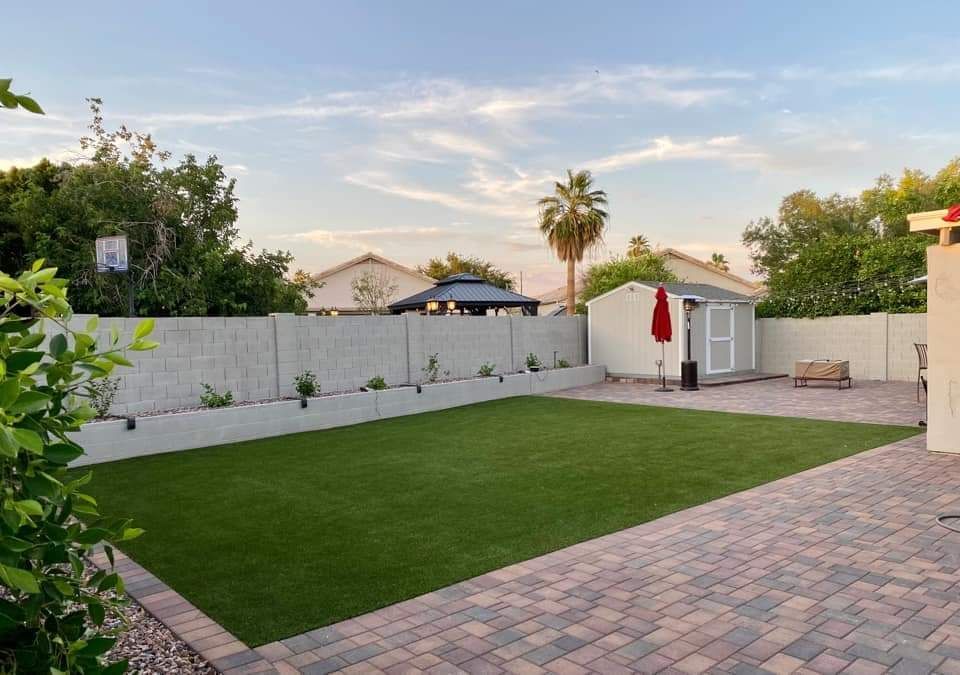 A backyard in Palm Valley FL with a well-manicured lawn and a paved patio area. There is a white storage shed in the right corner, a red umbrella, and some outdoor furniture on pristine Jacksonville Artificial Turf. The yard is enclosed by a white privacy fence, and there are trees and houses in the background.