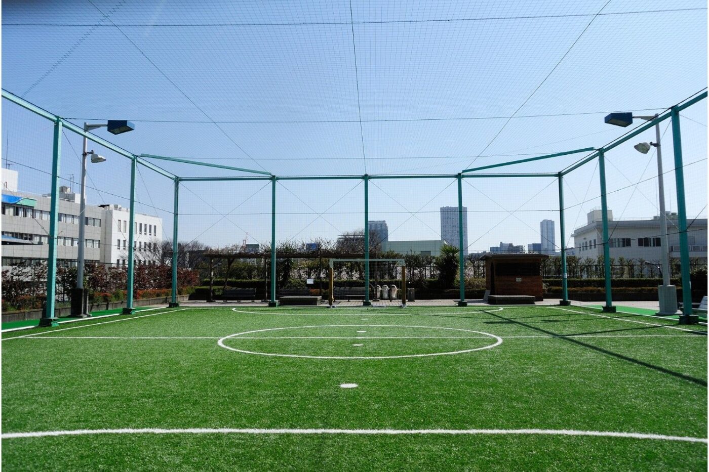 An outdoor rooftop soccer field with green artificial turf offers a dynamic outdoor living space. The field is enclosed by a net and green metal fencing, with buildings and skyscrapers towering in the background under a clear blue sky, showcasing expert fake turf installation.