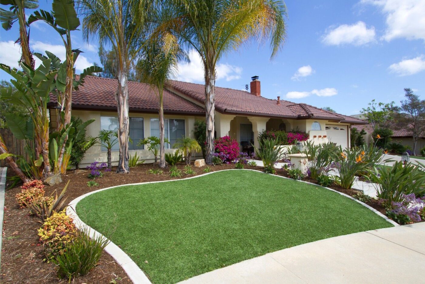 A single-story house with a red-tiled roof, palm trees, and a well-maintained front yard featuring lush green artificial grass, various plants, and flowers. A curved concrete driveway leads to a two-car garage. The sky is partly cloudy over Neptune Beach.