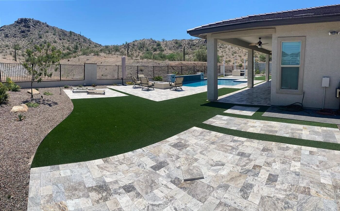 A backyard in Palm Valley, FL with a spacious patio featuring stone tiles, a pool under a covered area with a ceiling fan, lounge chairs by the pool, and Jacksonville Artificial Turf. The yard is enclosed by a fence, and the background showcases a hilly, desert landscape under a clear sky.