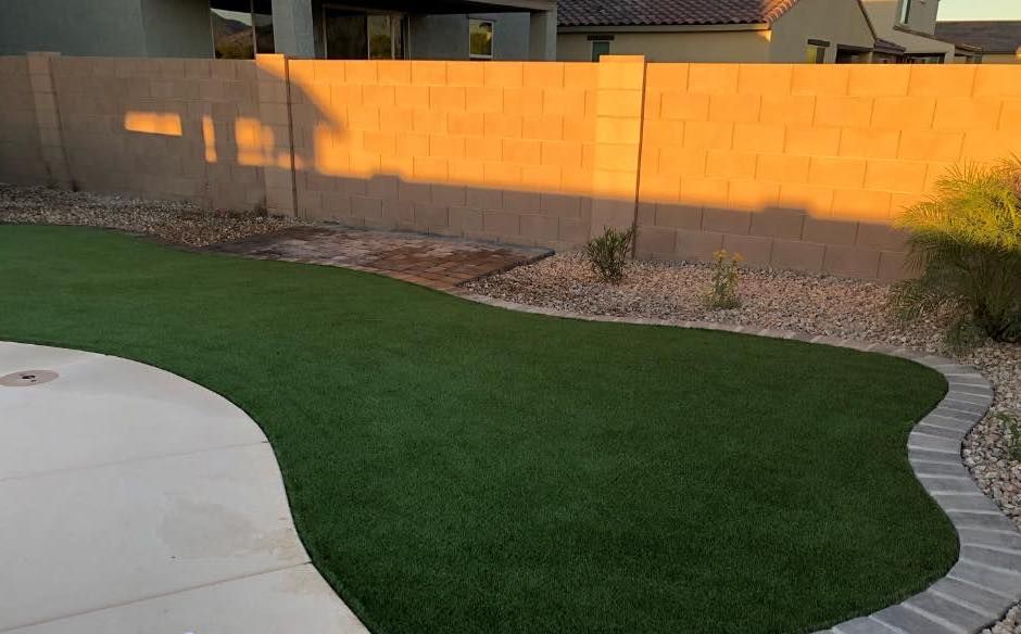 A house with a well-maintained front yard in Fernandina Beach boasts green grass, yellow flowering plants, and a curved stone border. A tree stands on the left side of the yard, while large potted plants enhance the outdoor living space atop a stone retaining wall in front of the house.