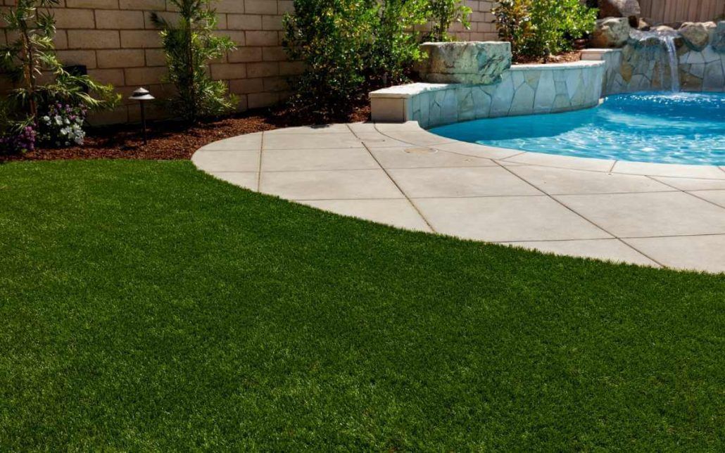 Close-up of a lawn with vibrant green grass. In the background, there are modern buildings and a black fence, slightly out of focus. The sky is clear and blue, suggesting a sunny day. The image captures the texture and detail of Jacksonville Artificial Turf. Get your free quote today!